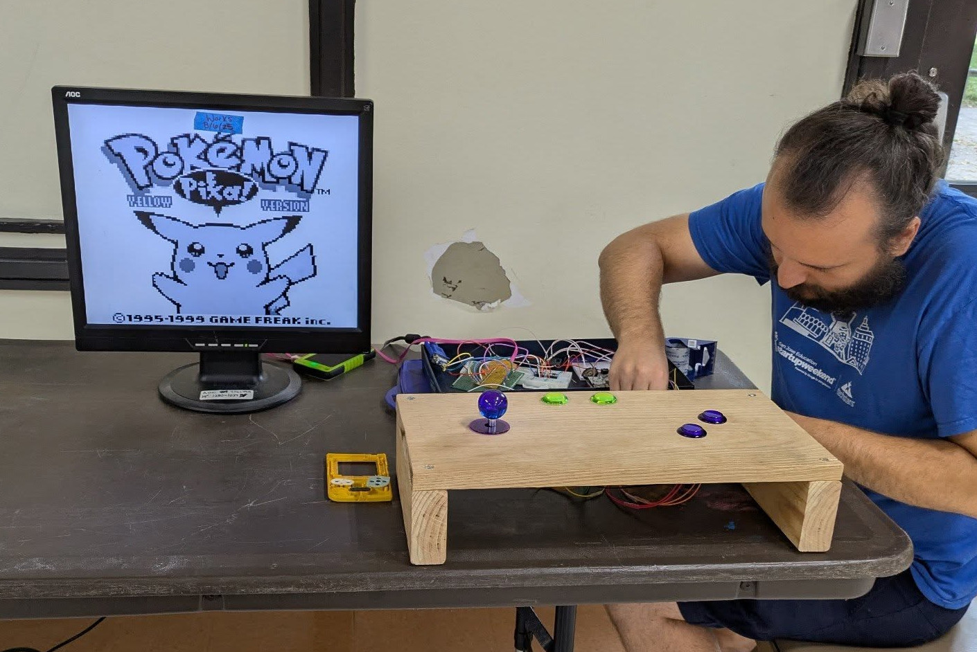 Jason fixing up wires on the giant arcade Gameboy, joystick and big arcade buttons for controls and Pokemon yellow showing on the computer monitor used as a screen.
